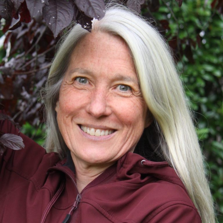 Barbara Treffert Frau mit langen, blonden Haaren, die lächelt und vor grünen Pflanzen steht.